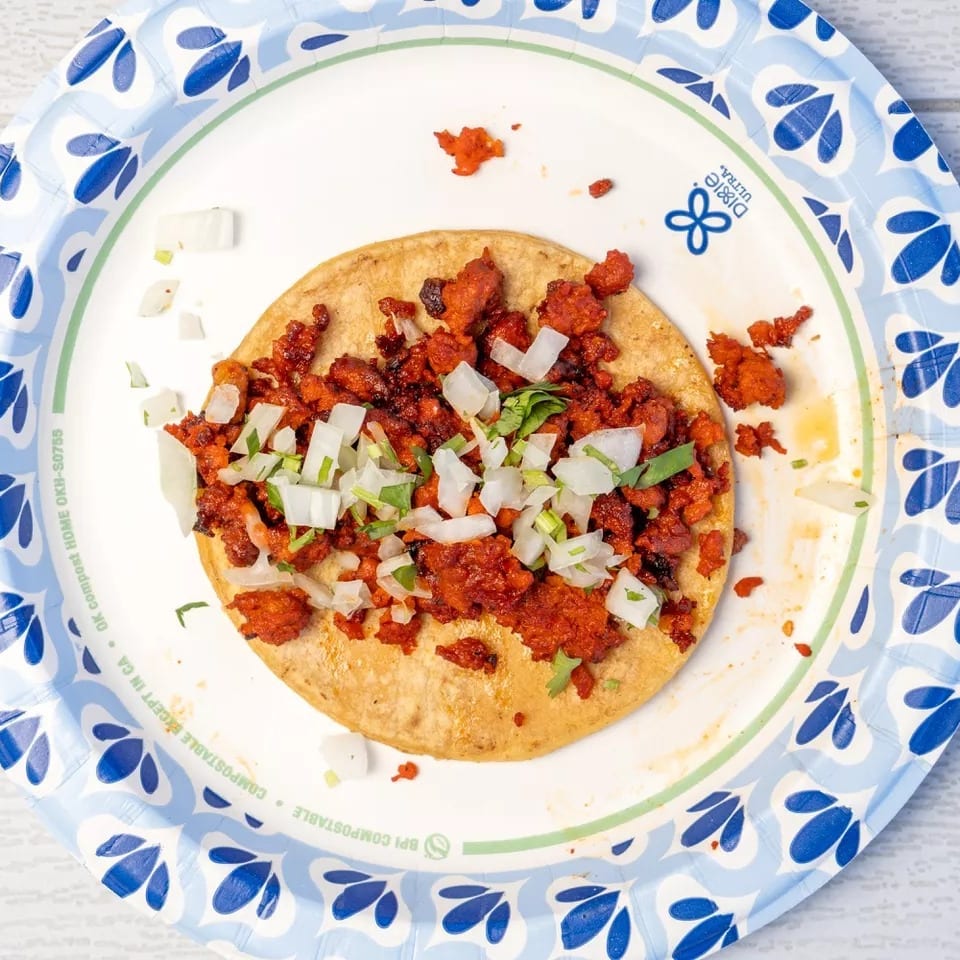 Al pastor taco with chopped onions and cilantro on a blue and white paper plate.