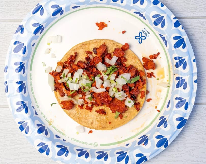 Al pastor taco with chopped onions and cilantro on a blue and white paper plate.