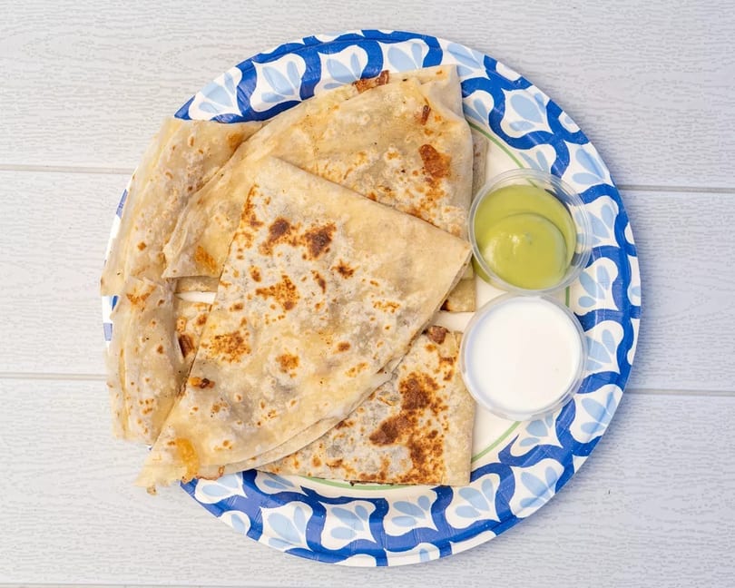 Golden-brown quesadillas on a blue and white paper plate with green and white dipping sauces.