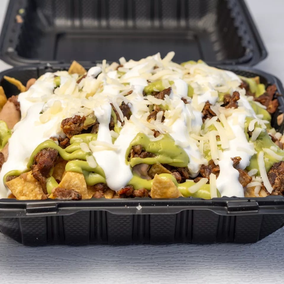 Loaded nachos with meat, guacamole, sour cream, and shredded cheese in a black takeout container.