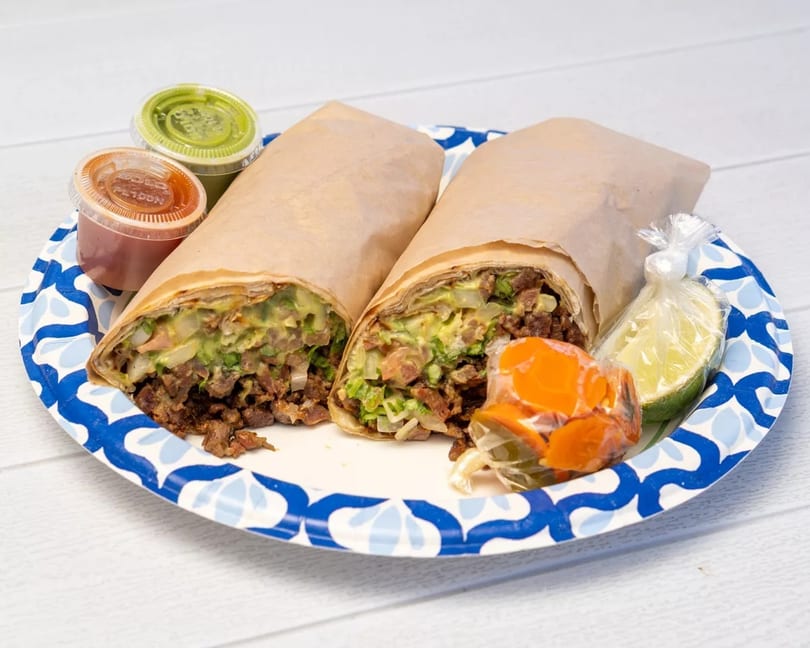 Two large burritos, unwrapped to show meat and guacamole, with salsa, lime, and carrots on a plate.