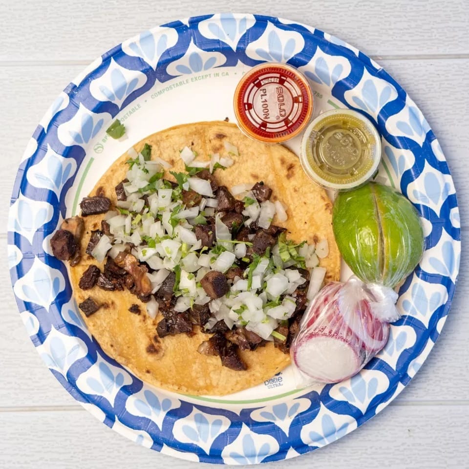 Overhead view of a taco with meat, onions, and cilantro, with lime and sauces on a paper plate.