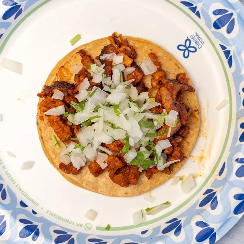 Al pastor taco on a corn tortilla, topped with diced onions and cilantro, on a paper plate.