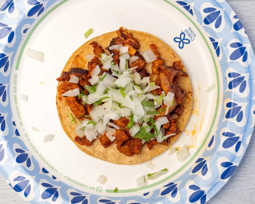 Al pastor taco on a corn tortilla, topped with diced onions and cilantro, on a paper plate.