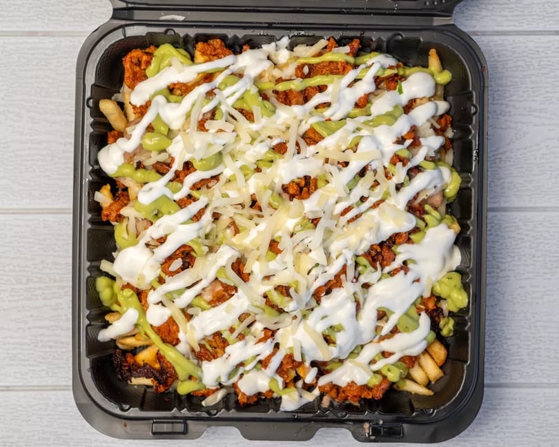 Loaded fries with chorizo, white and green sauces, and shredded cheese in a black takeout container.