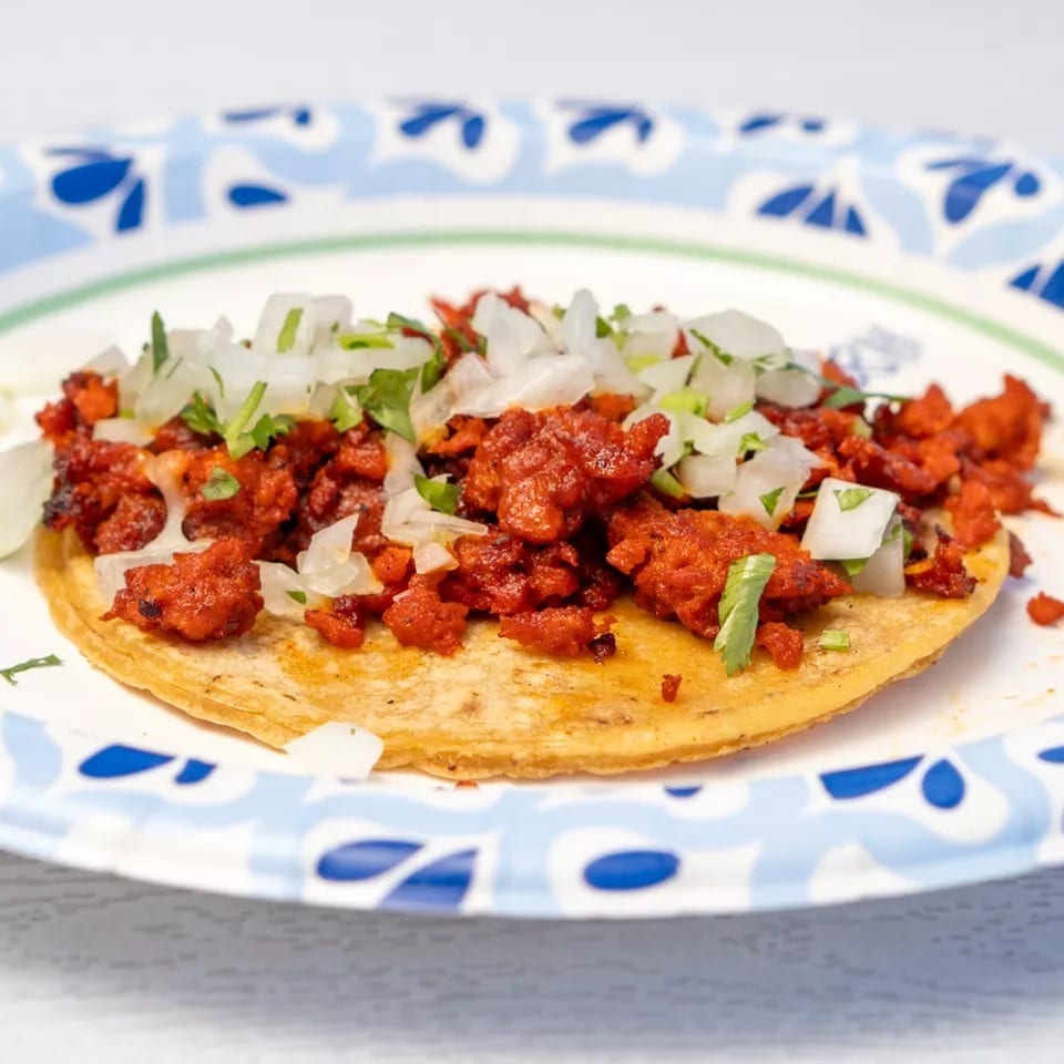 Al pastor taco with red marinated pork, diced onions, and cilantro on a paper plate.