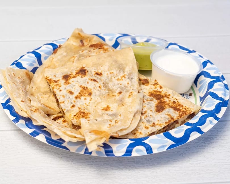 Quesadillas on a blue and white paper plate with sides of guacamole and sour cream.