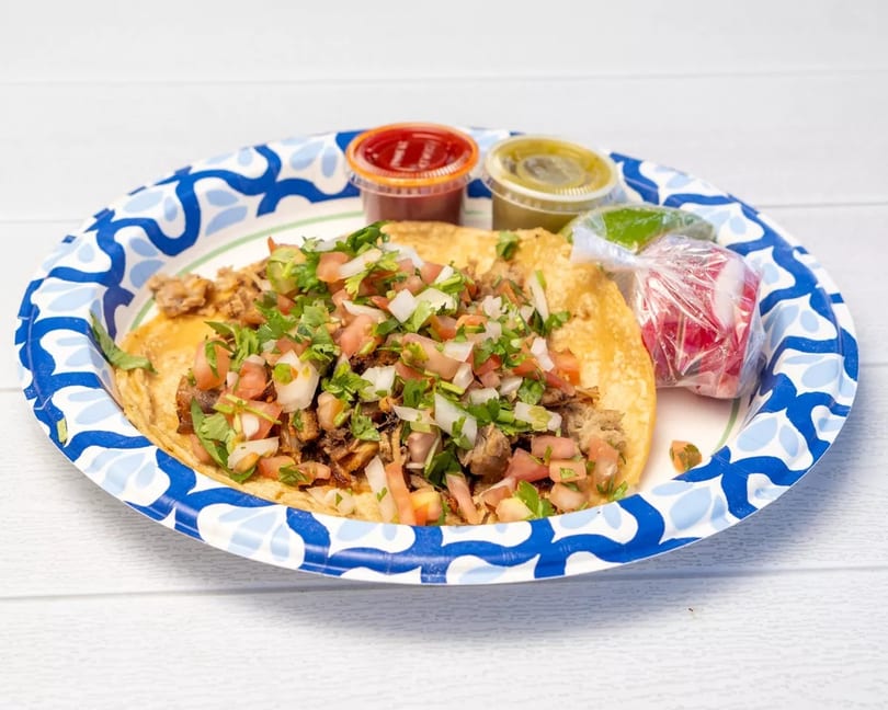 Two tacos with meat, pico de gallo, and cilantro on a paper plate with sauces and radishes.