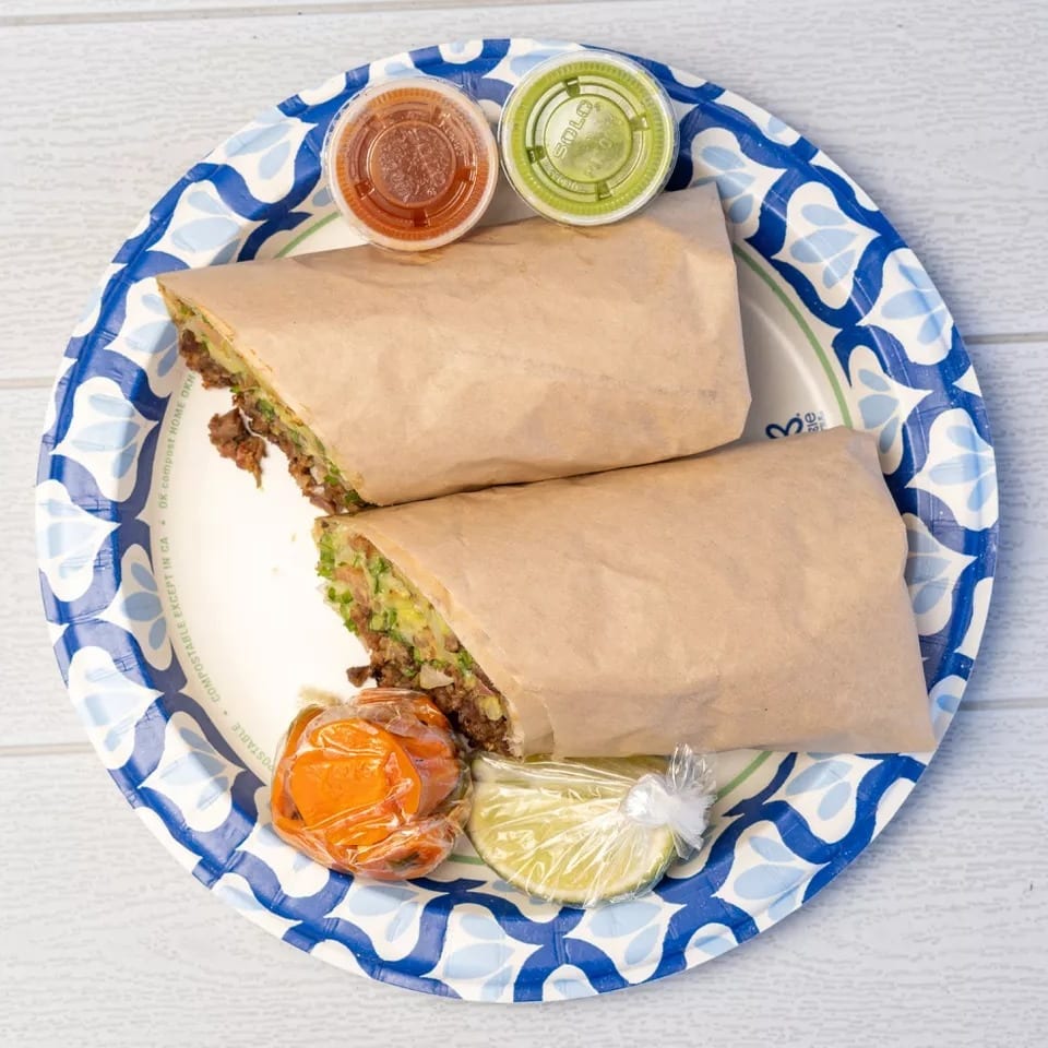 Two burritos wrapped in paper, with salsa, guacamole, and lime wedges on a blue and white plate.