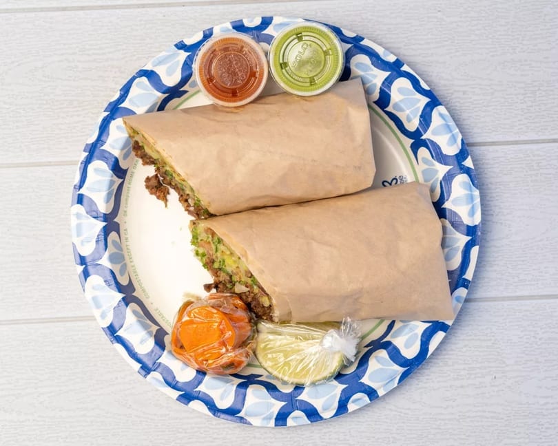 Two burritos wrapped in paper, with salsa, guacamole, and lime wedges on a blue and white plate.