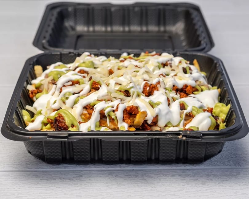 Loaded fries with chorizo, shredded cheese, sour cream, and guacamole in a black takeout container.