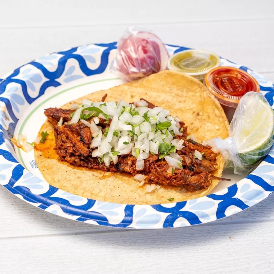 A single taco with meat, onions, and cilantro, served with lime and sauces on a paper plate.