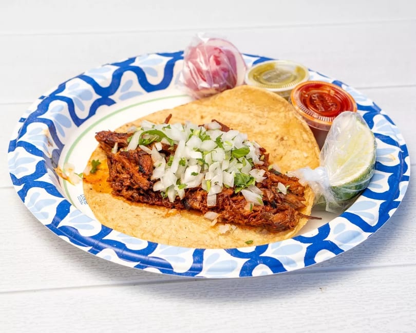 A single taco with meat, onions, and cilantro, served with lime and sauces on a paper plate.