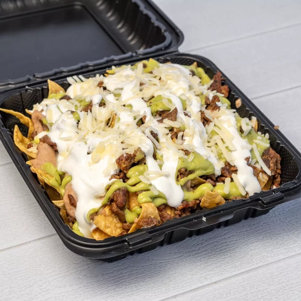 Nachos with meat, guacamole, sour cream, and shredded cheese in a black takeout container.