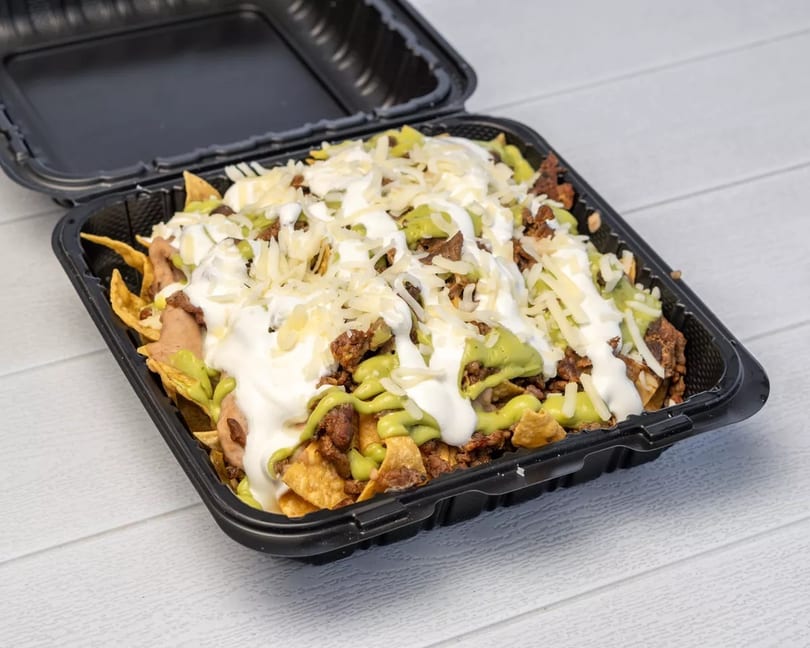 Nachos with meat, guacamole, sour cream, and shredded cheese in a black takeout container.
