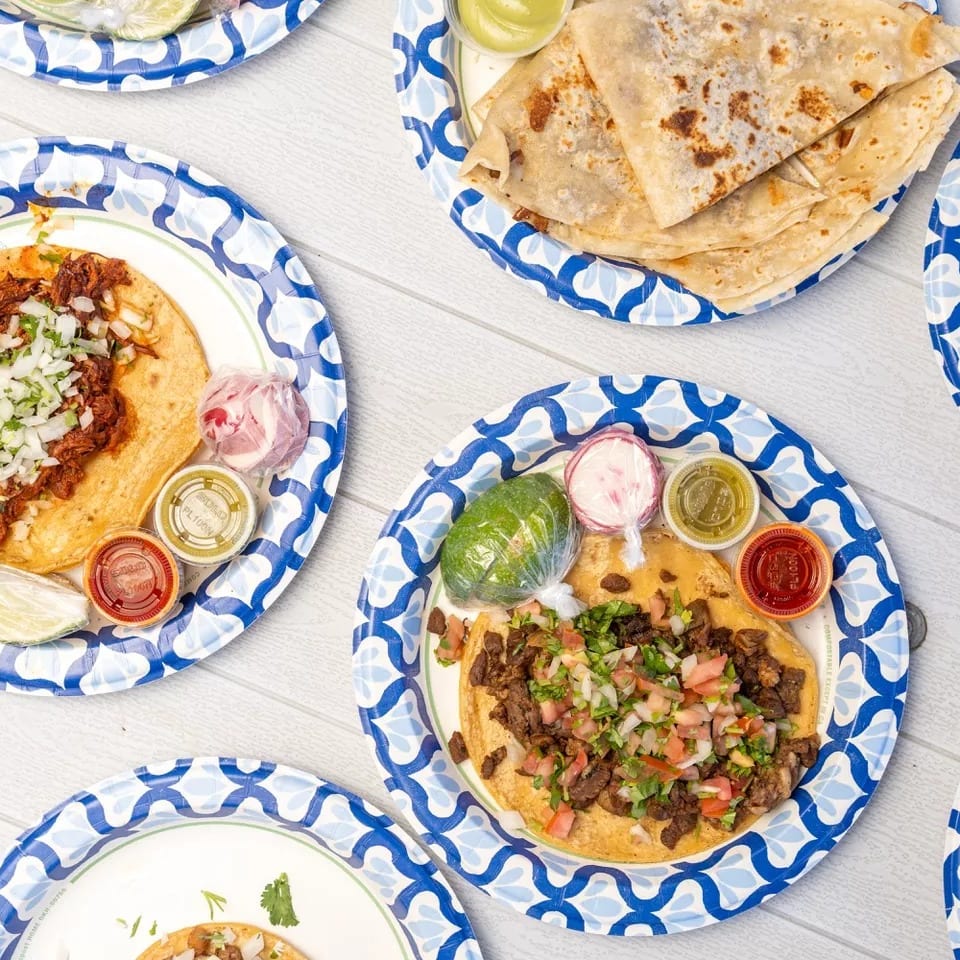 Several plates of tacos and quesadillas with various toppings and sauces on a white wooden table.
